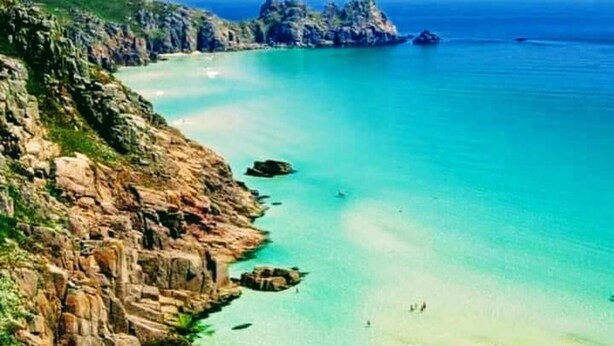 A coastal landscape with a clear blue sea, rocky cliffs, and a sandy beach.