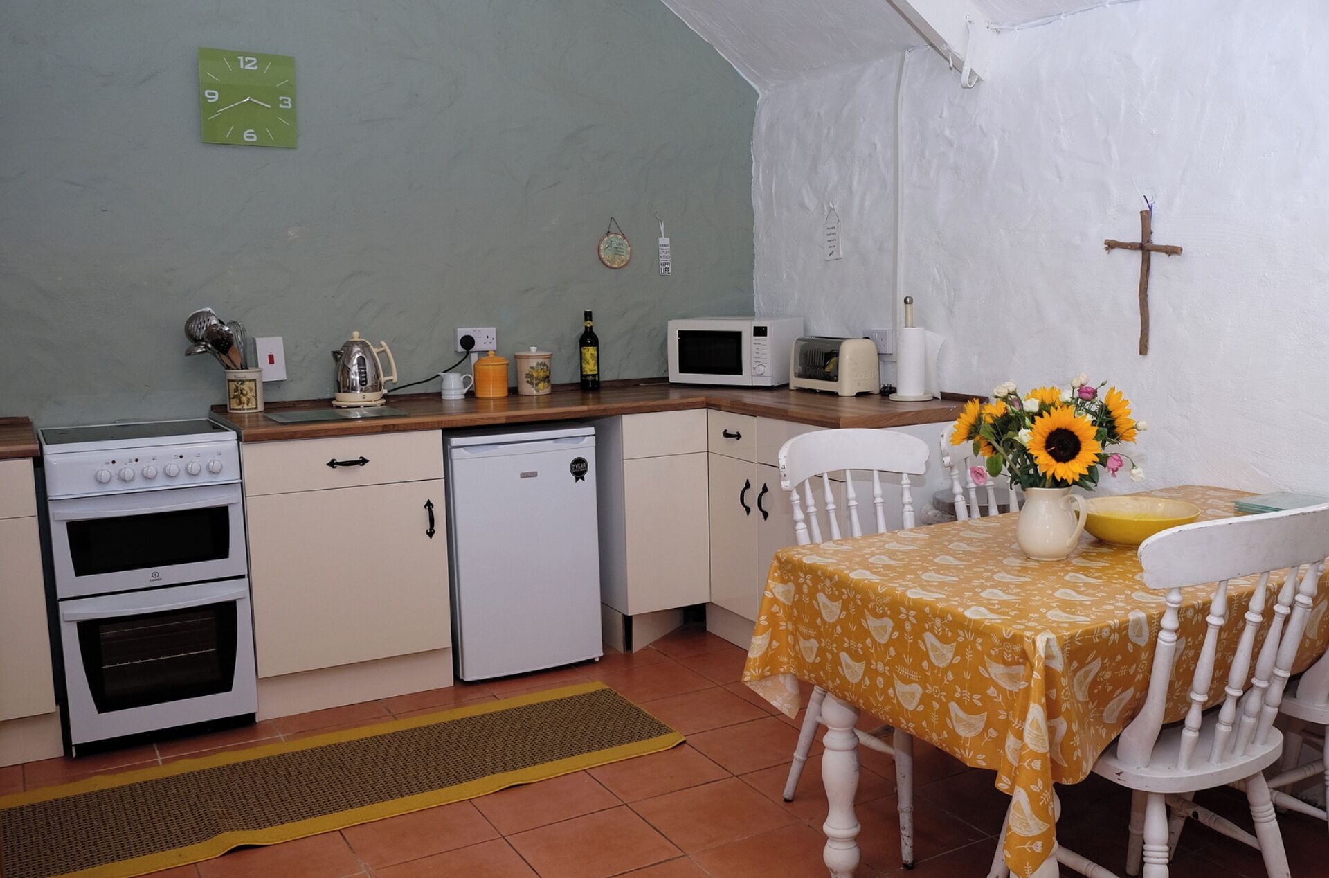A compact kitchen with a stove, microwave, and small refrigerator. There's a dining table with a yellow tablecloth and a vase of sunflowers.