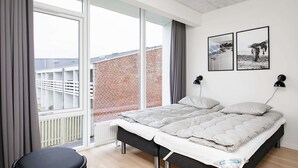 A modern bedroom with a large bed, a chair, and a view of a brick building through a glass door. - 5 Star Holiday Home in Ringkobing (Ringkobing)