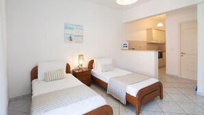 A hotel room with two single beds, a nightstand with a lamp, a painting on the wall, and a kitchenette in the background. - Irini studio for 2, central location and 5 minutes walk from the sandy beach (Corfu)