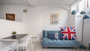 A modern living space with a white brick wall, a blue sofa with a Union Jack pillow, a dining table with chairs, and a small plant on the table. - Framery 3 - Funky studio in trendy Shoreditch (London)