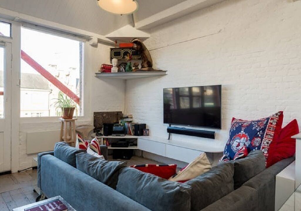 A living room with a sofa, a flat-screen TV mounted on the wall, a shelf with various items, and a window with a view of buildings.