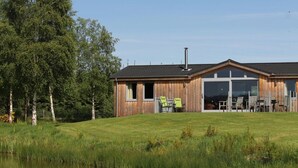 A wooden cabin with a large glass door and windows, situated next to a body of water with a grassy field and trees in the background. - Exceptional lodge in private woodland with luxury steam room (Moneydie)
