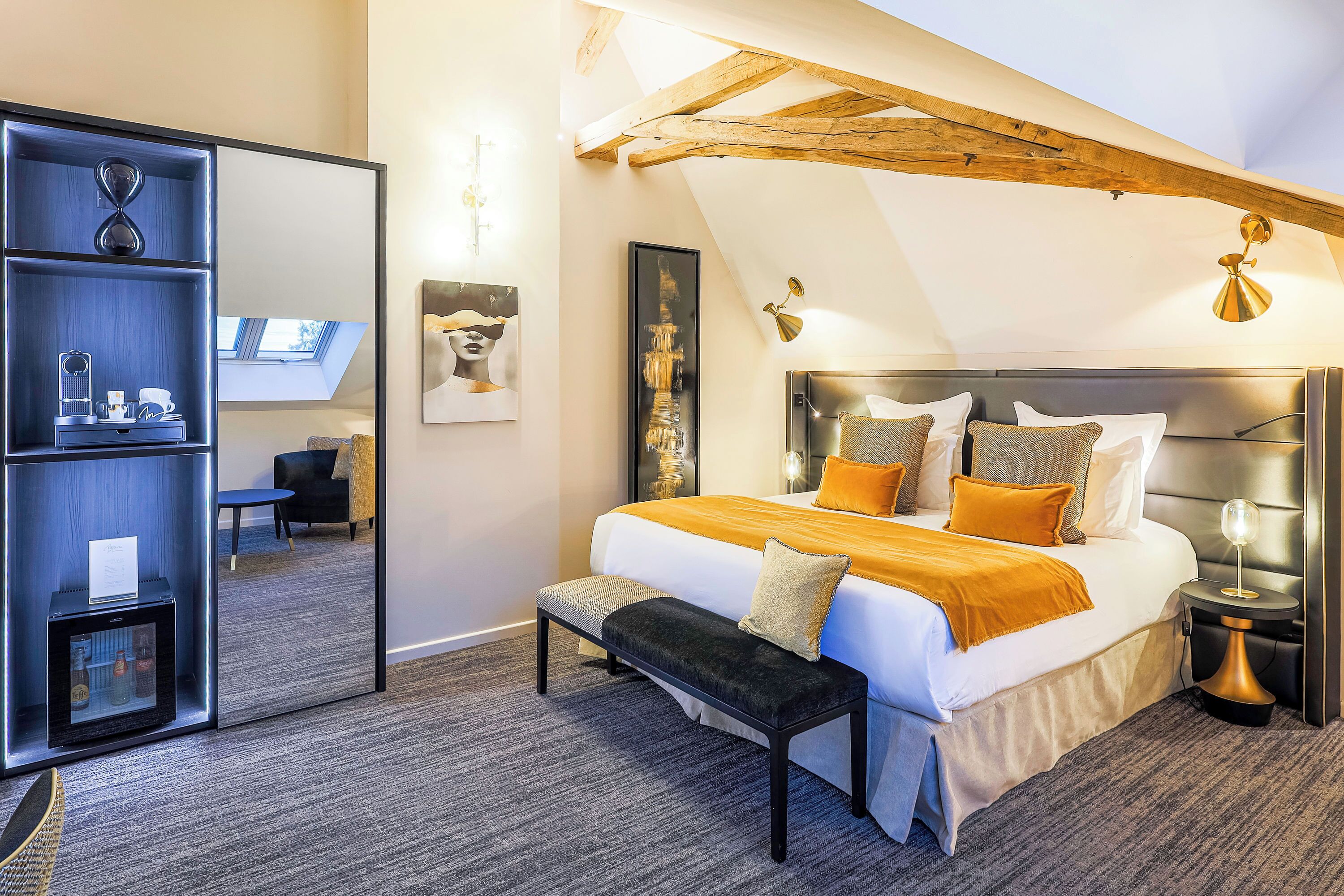A modern bedroom with a large bed, a bench, a mirror, and a small table with a lamp.