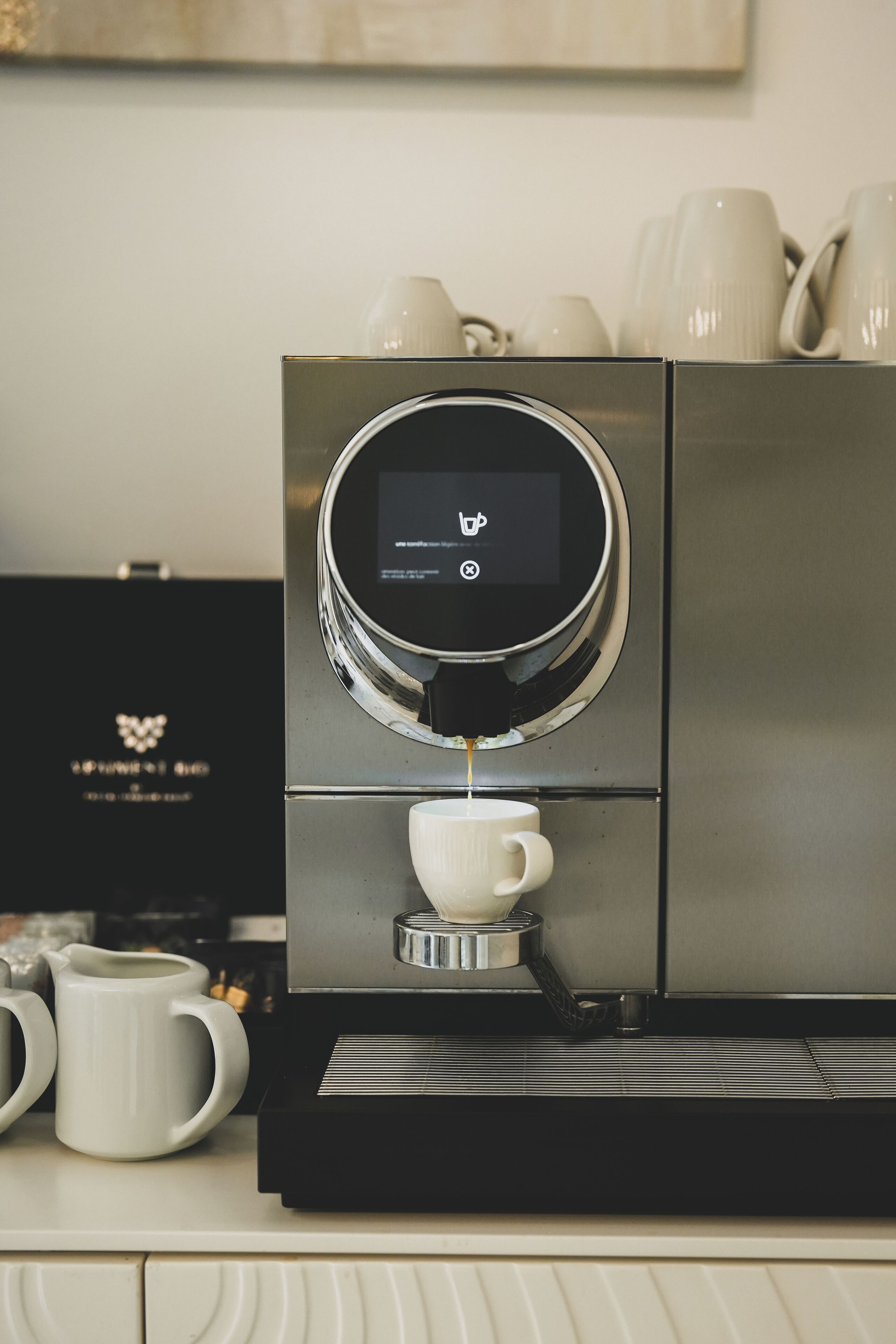 A coffee machine with a digital interface, a coffee cup, and a set of white mugs on a counter.