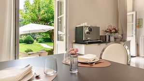 Un coin repas avec une table dressée pour deux personnes, une machine à café et une vue sur un jardin par de grandes fenêtres ouvertes.