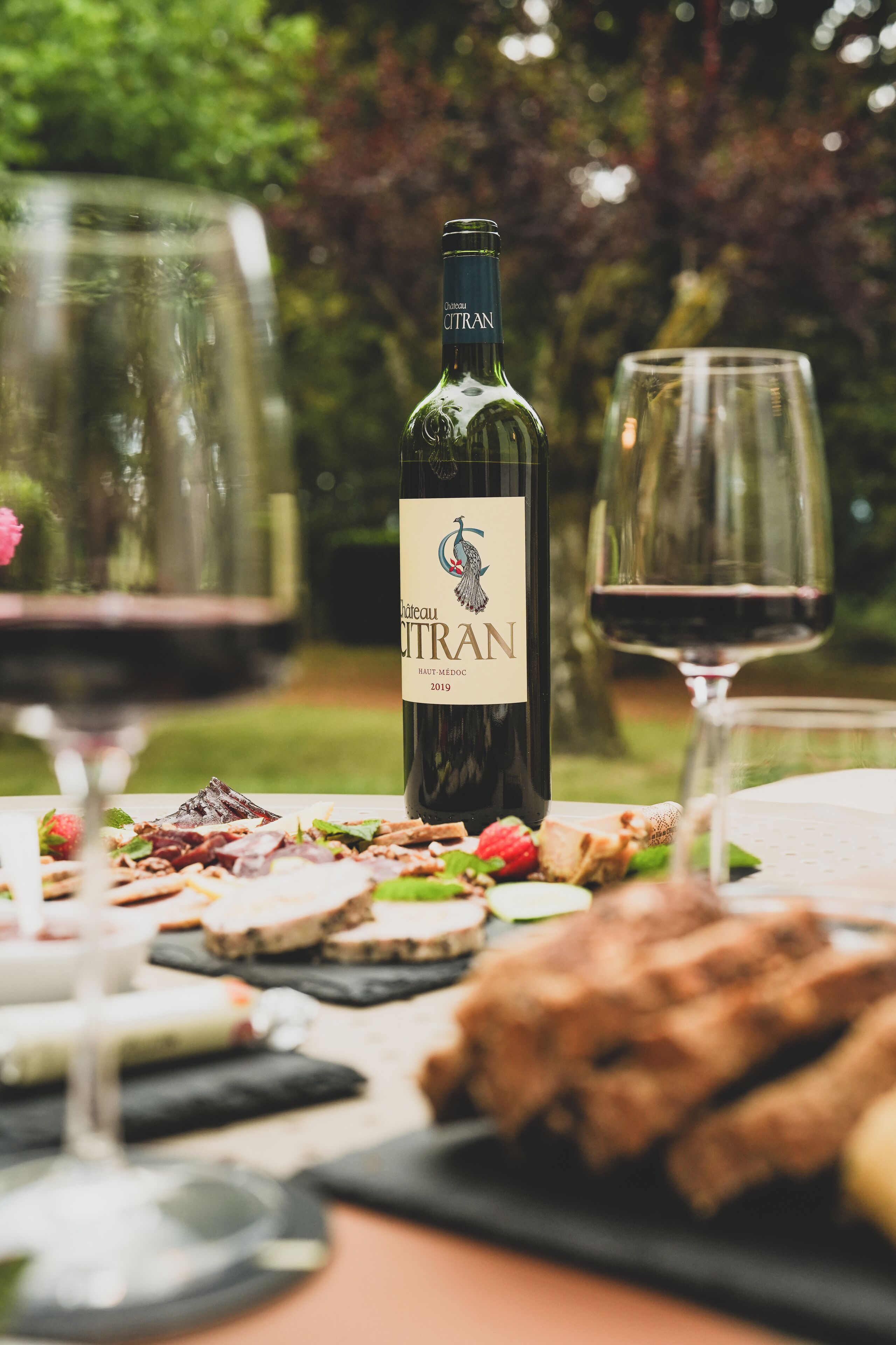 A bottle of Chateau Cortan wine, a glass of red wine, and a plate with various cheeses and crackers on an outdoor table.