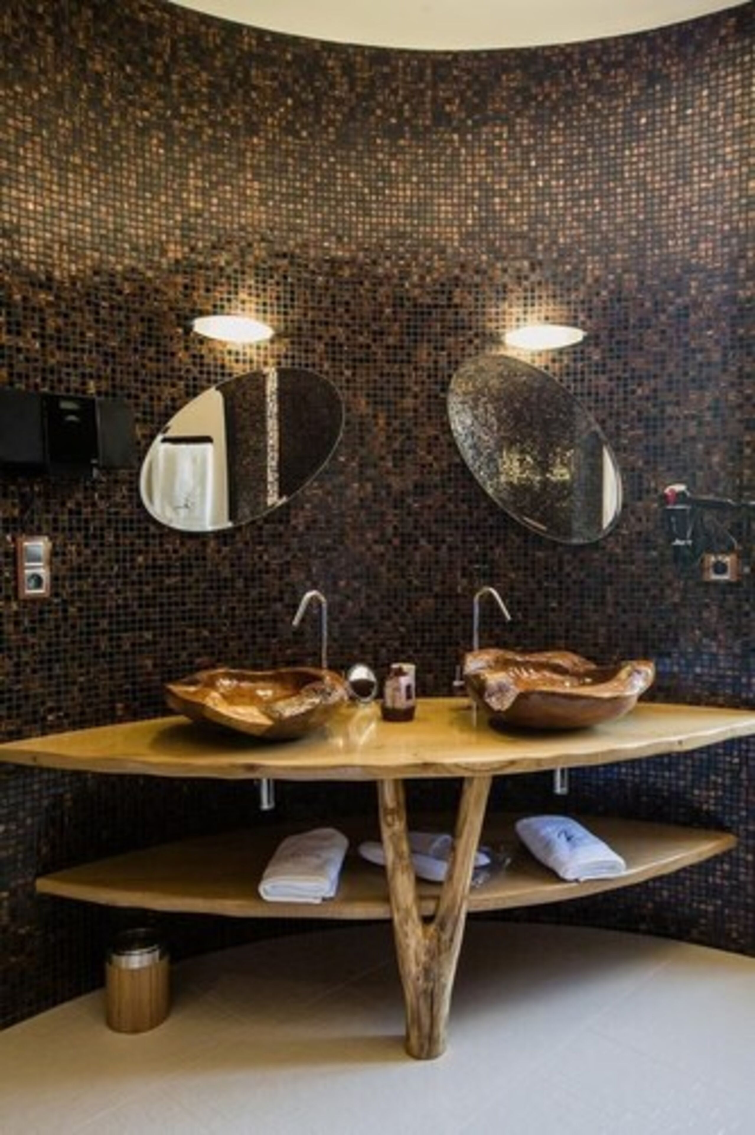 Une salle de bain avec un mur décoré de mosaïques, deux miroirs ronds, un meuble-lavabo en bois avec un vasque et une étagère avec des serviettes.