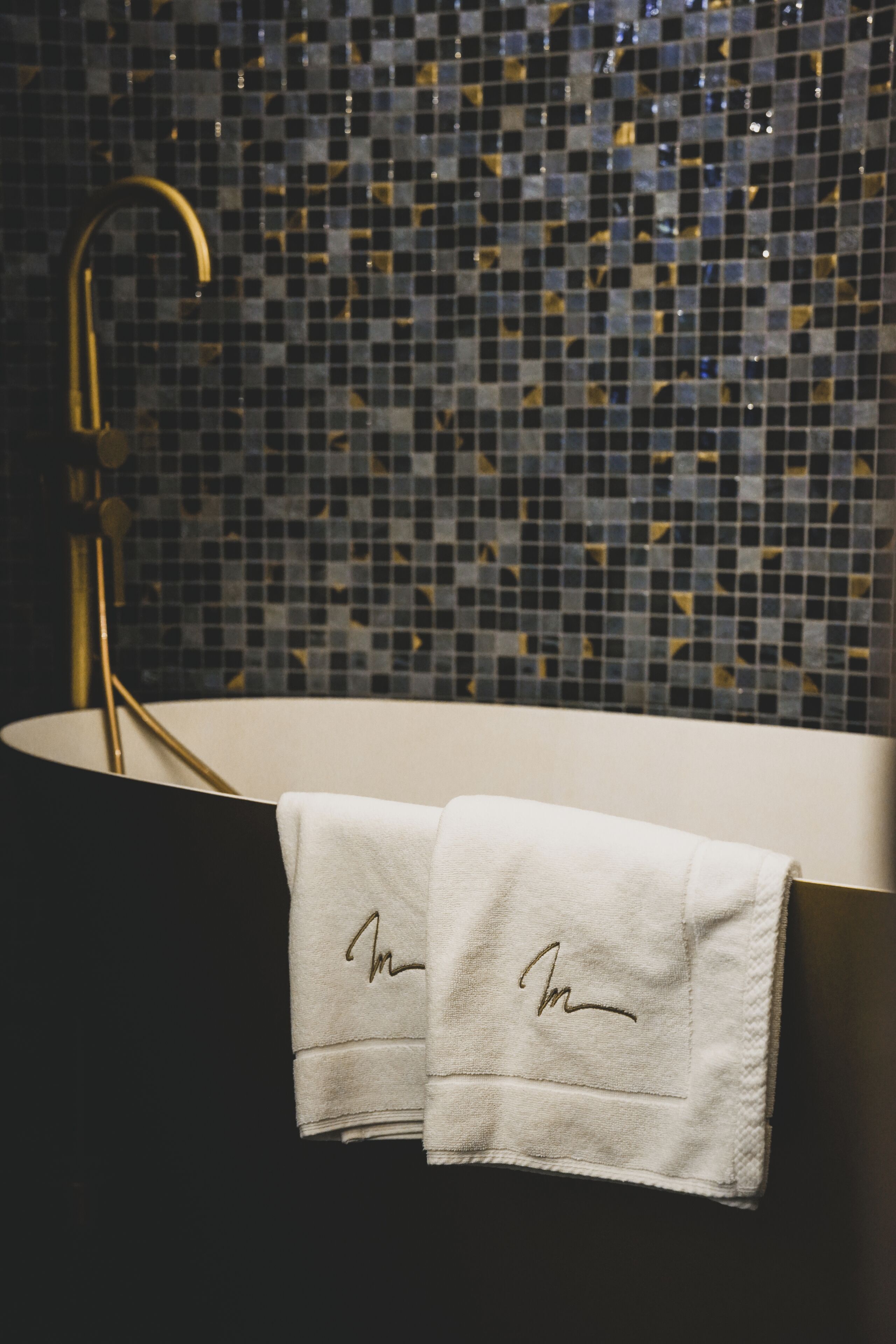 Ein Badezimmer mit einem schwarzen Waschbecken, einem goldenen Wasserhahn und einer Wand mit Mosaikfliesen. Zwei mit Unterschriften versehene Handtücher hängen an der Wand.