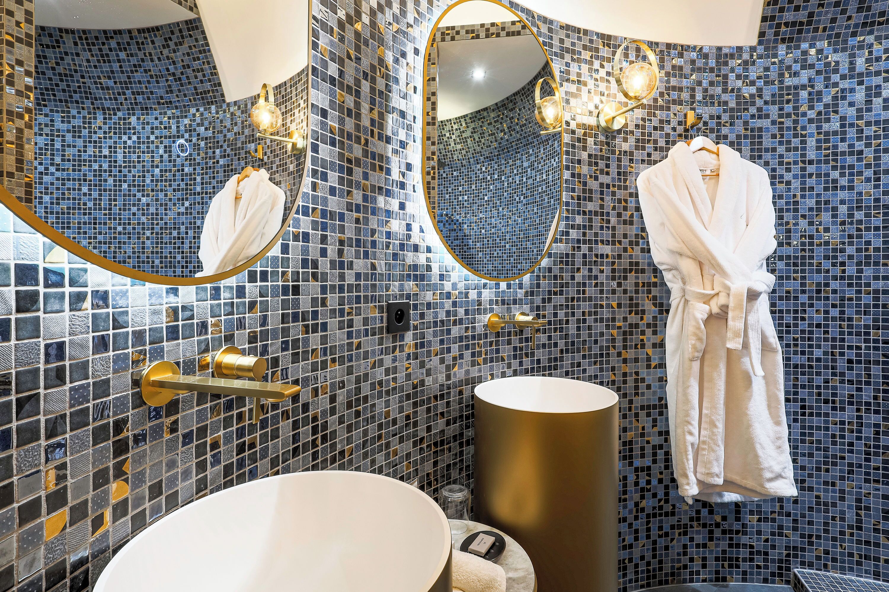 Un bagno con parete in mosaico, uno specchio rotondo, una vasca da bagno bianca e un accappatoio appeso al muro.