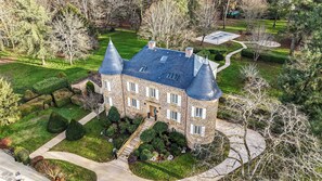 Une vaste demeure en pierre, dotée d’un toit à forte pente et de nombreuses cheminées, entourée de jardins magnifiquement entretenus et d’une allée sinueuse.