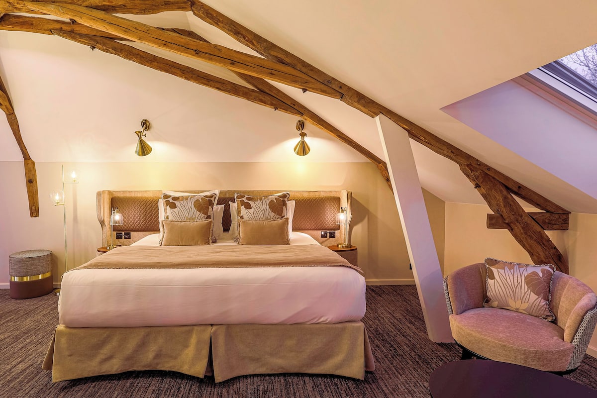 A bedroom with a large bed, wooden beams, a skylight, and a chair.