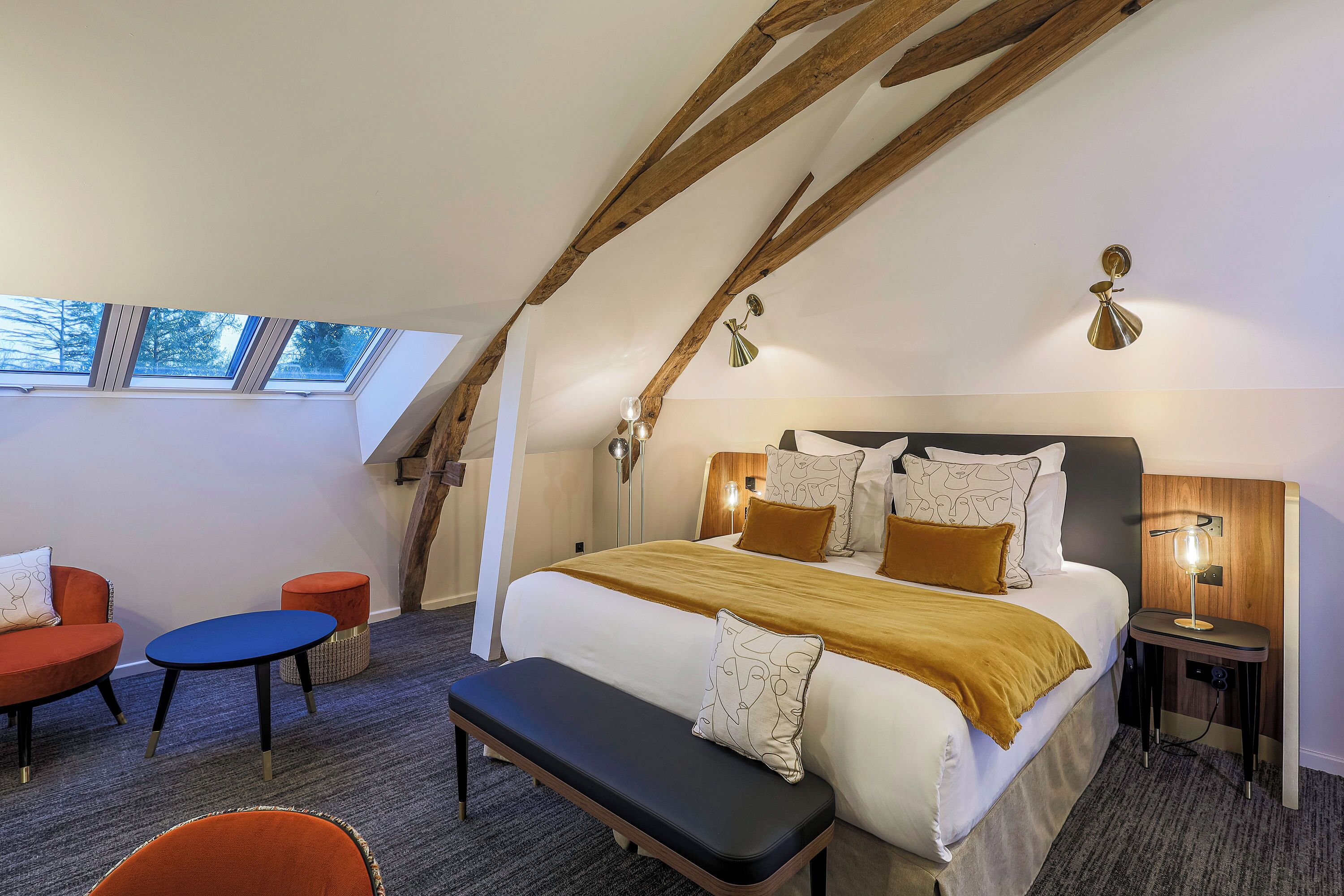 A bedroom with a sloped ceiling, a large bed with multiple pillows, a bench, a small table, and a chair. There are two bedside tables with lamps.