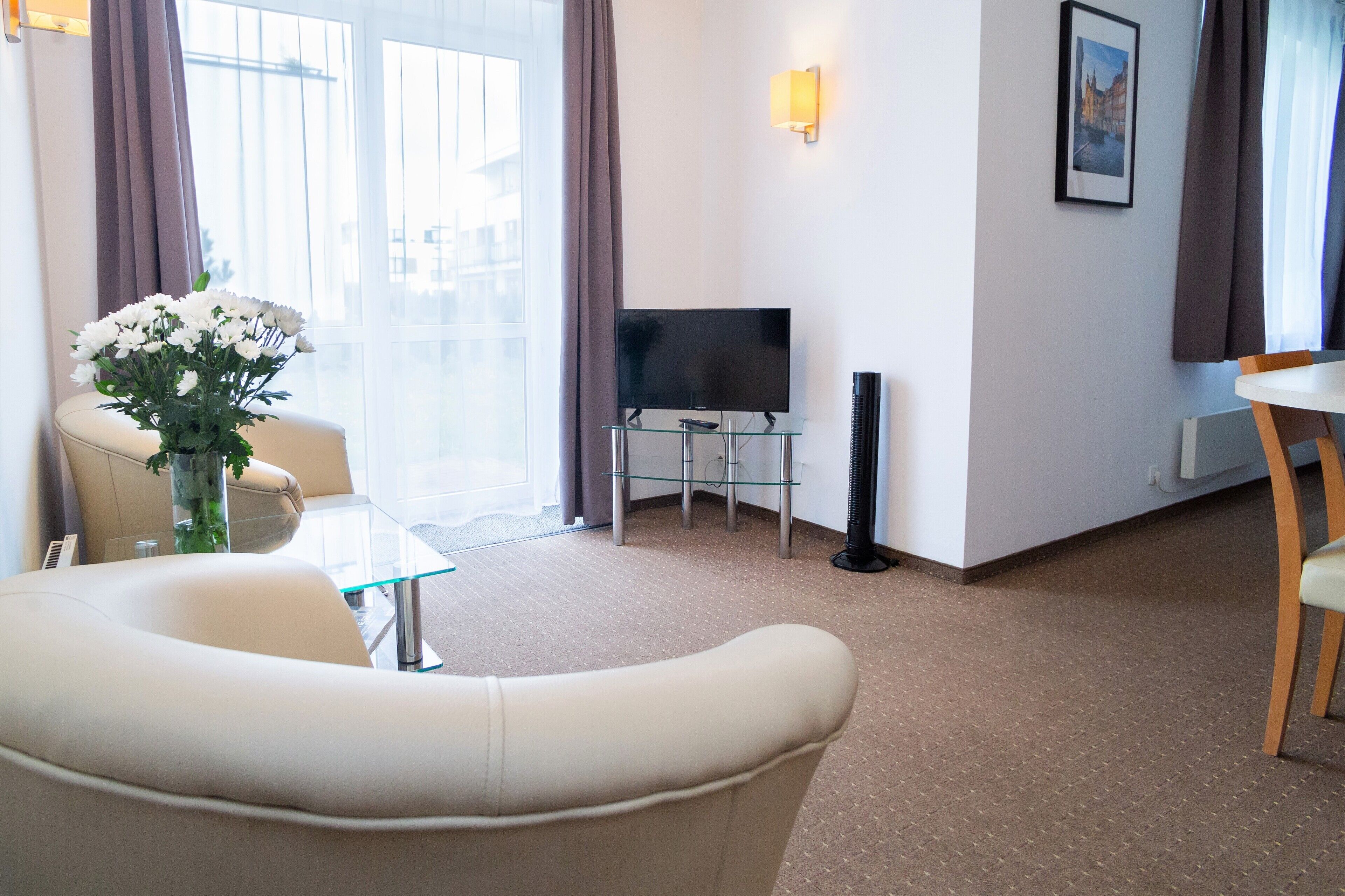 A hotel room with a sofa, a glass table, a flat-screen TV, a vase with flowers, and a picture on the wall.