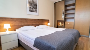 Un lit bien fait, recouvert d’une couverture grise, avec deux lampes de chevet, une tête de lit en bois, un tableau représentant un paysage urbain encadré au mur et une armoire en bois dotée d’un miroir.