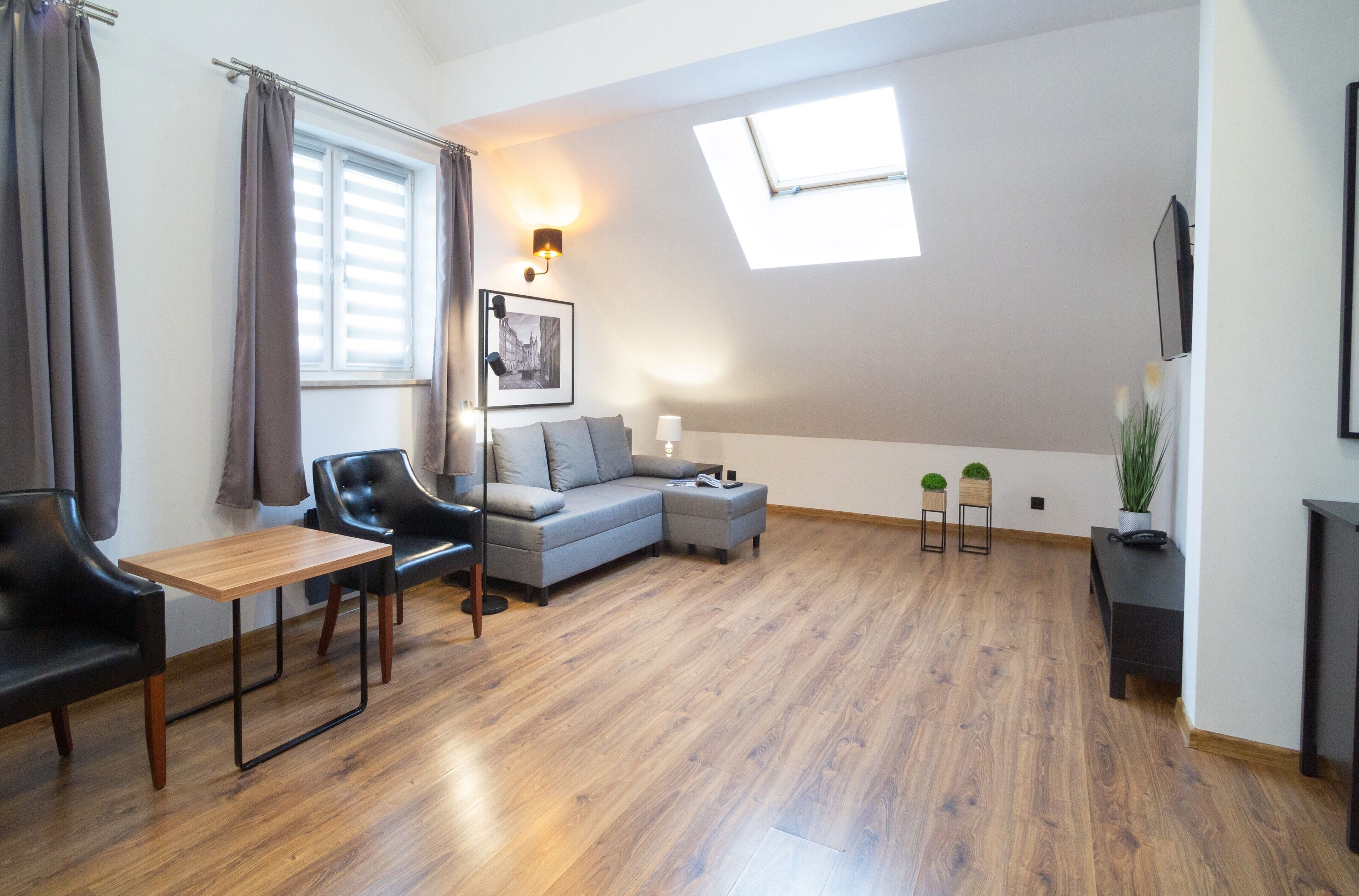 A modern living room with a wooden floor, a grey sofa, a black leather chair, a small wooden table, and a skylight.