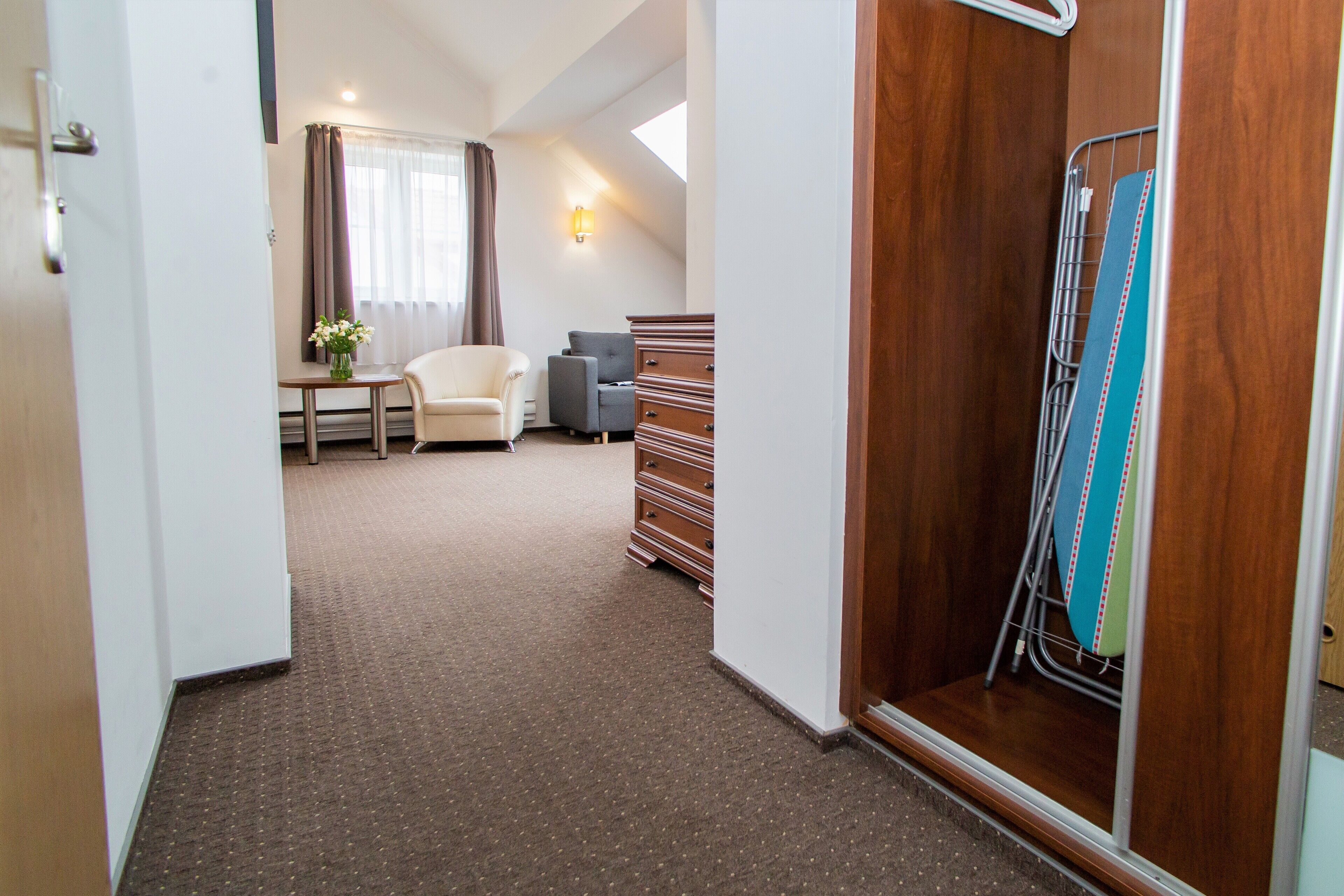 A hotel room with a carpeted floor, a bed, a dresser, a chair, a small table, and a window with curtains.