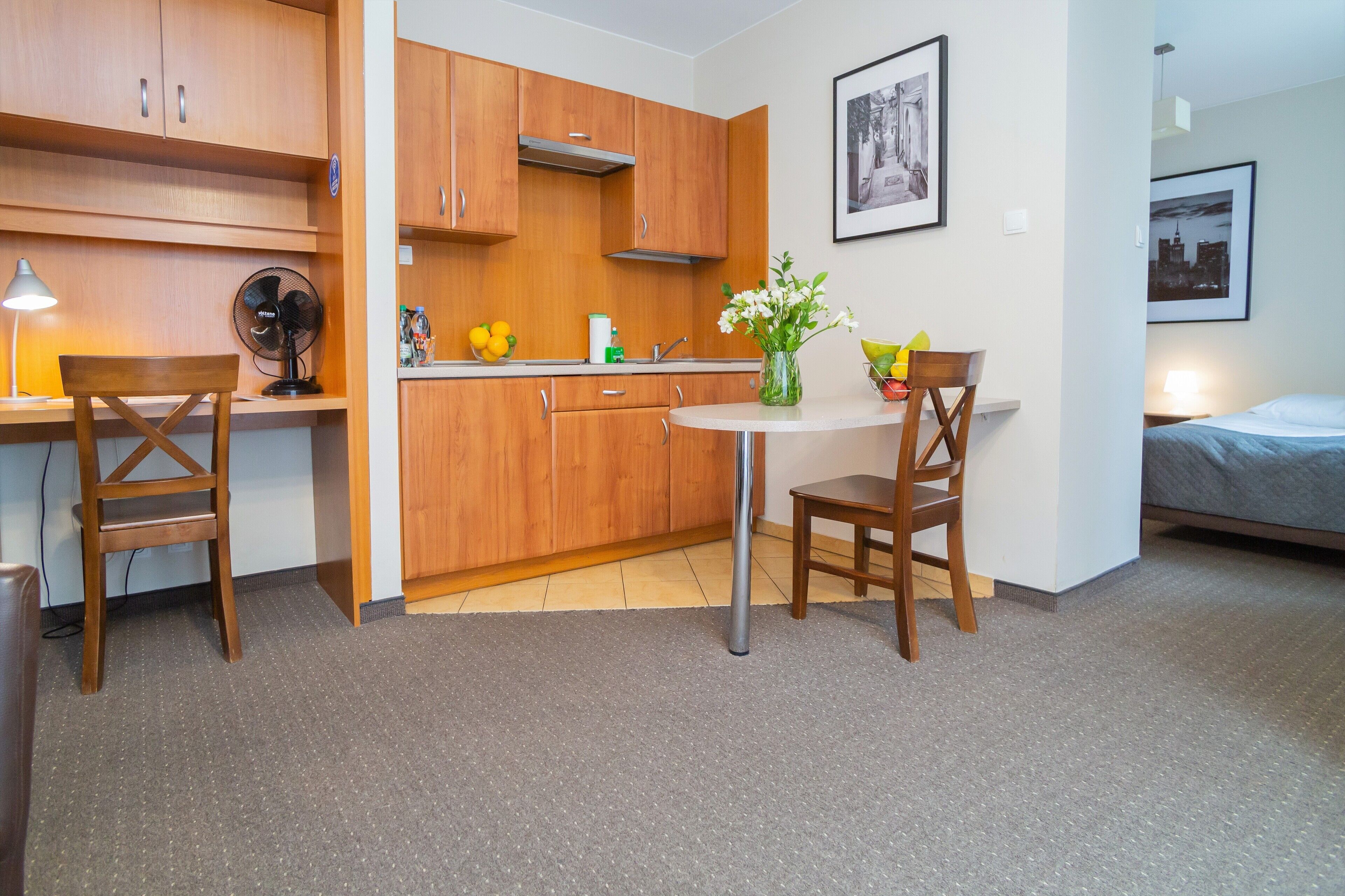 A compact kitchen with wooden cabinets, a dining table with chairs, a small round table with a vase, and a bed in the adjacent room.