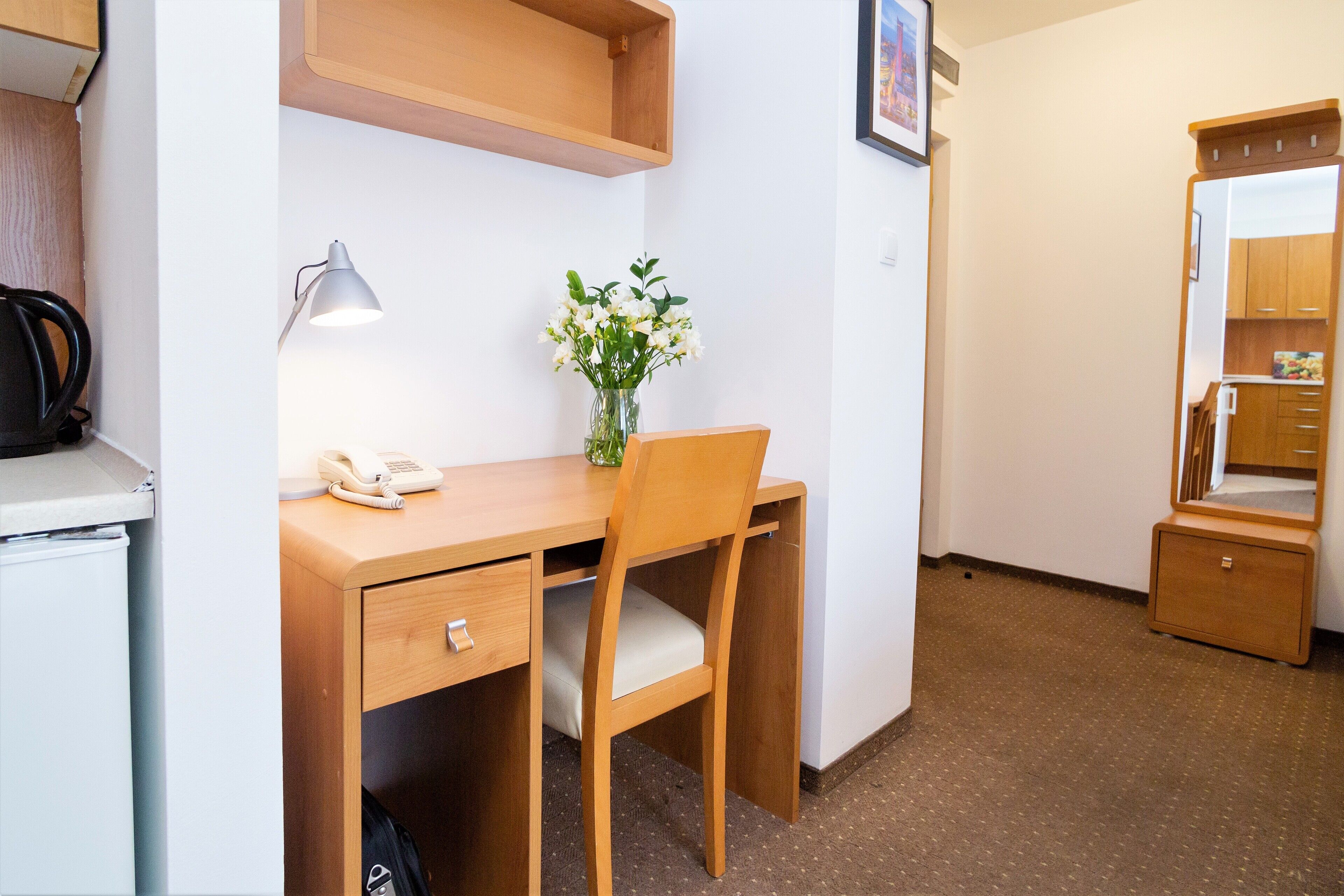 A compact hotel room with a wooden desk, a chair, a small bedside table, a mirror, and a wall-mounted shelf.
