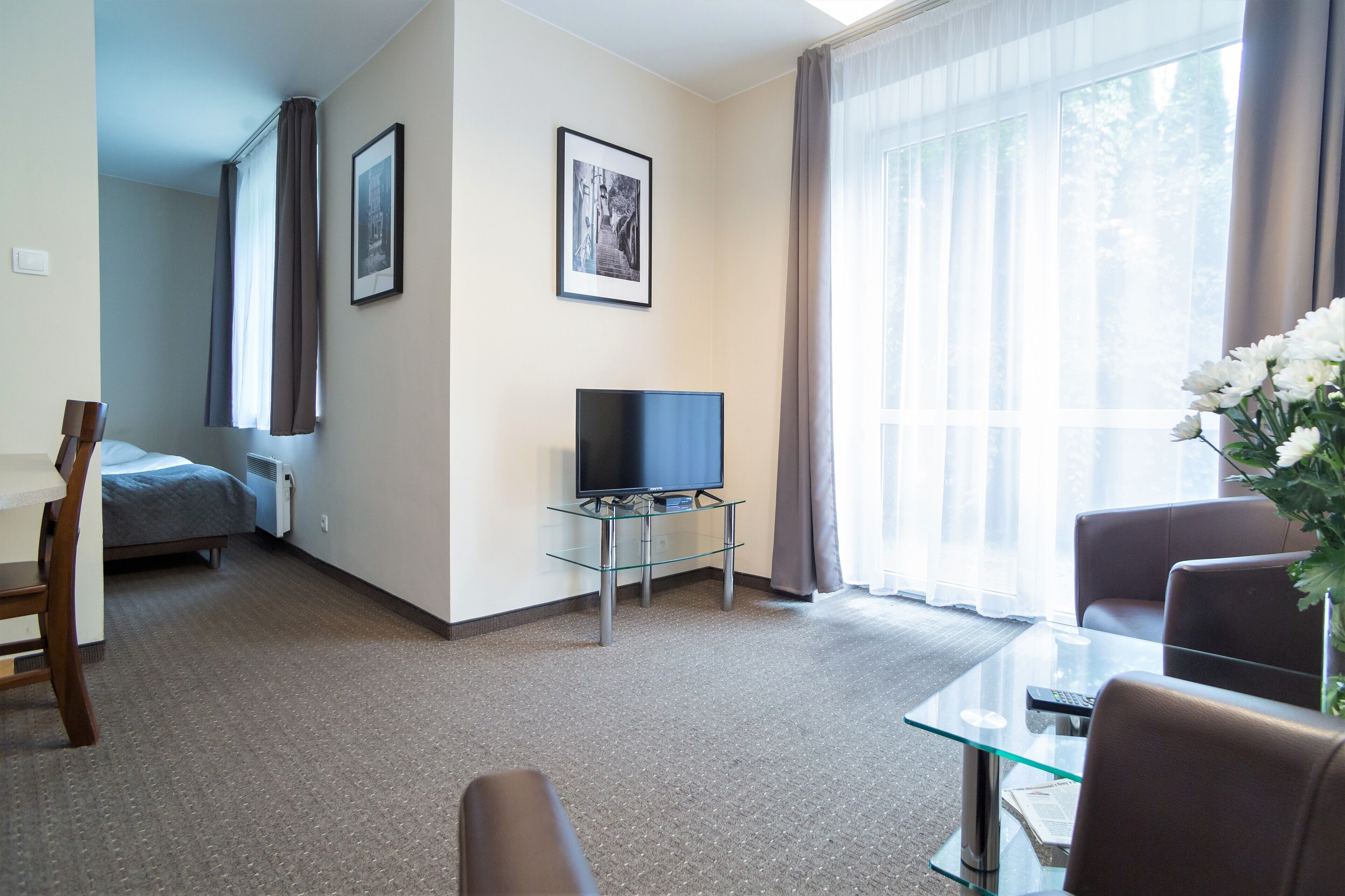 A hotel room with a bed, a TV on a glass table, a seating area with a table, and a window with curtains.