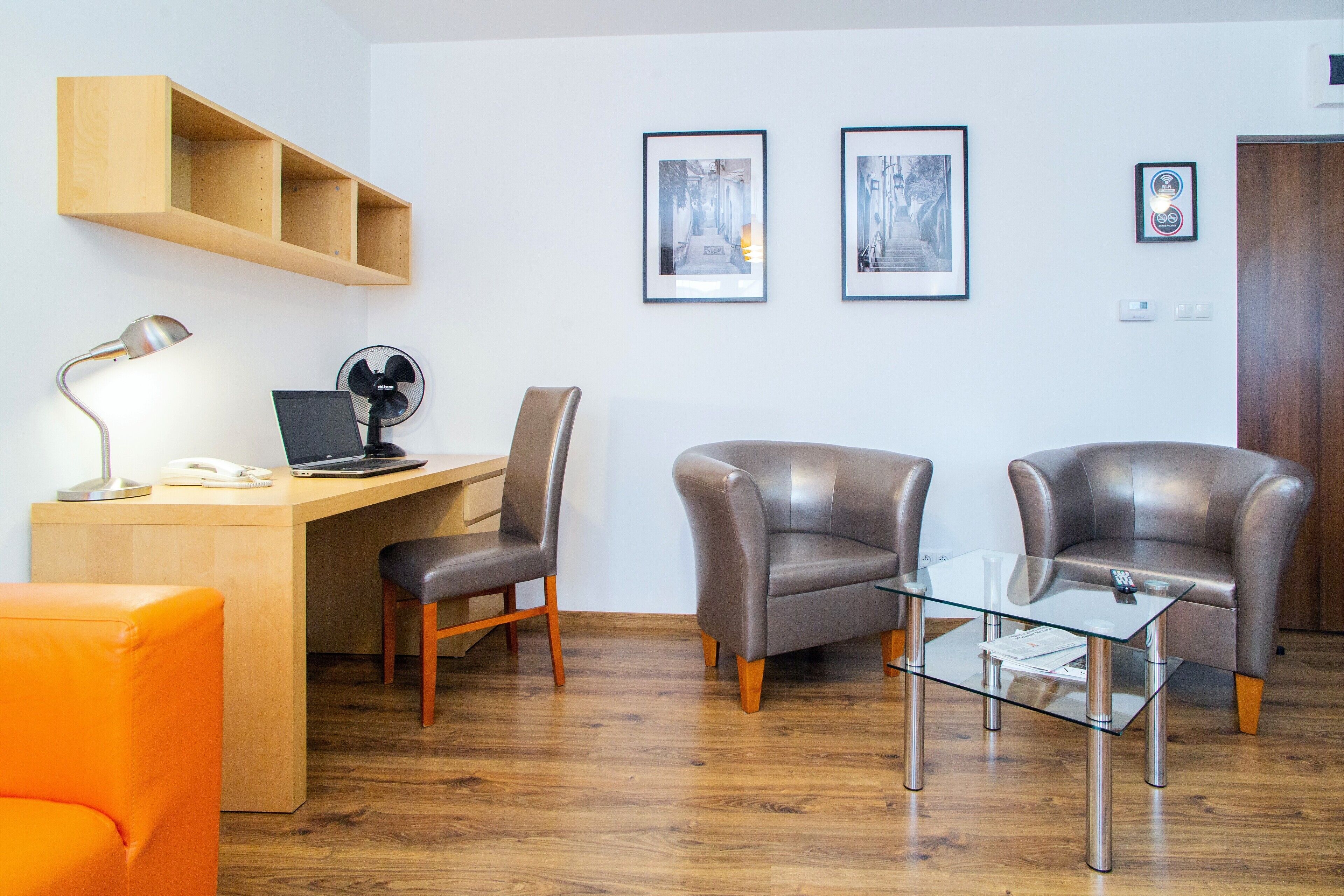 Una sala moderna con un escritorio de madera, dos sillones de cuero, una mesa de centro de vidrio y un sofá naranja brillante. Hay cuadros enmarcados en la pared y una estantería de madera sobre el escritorio.