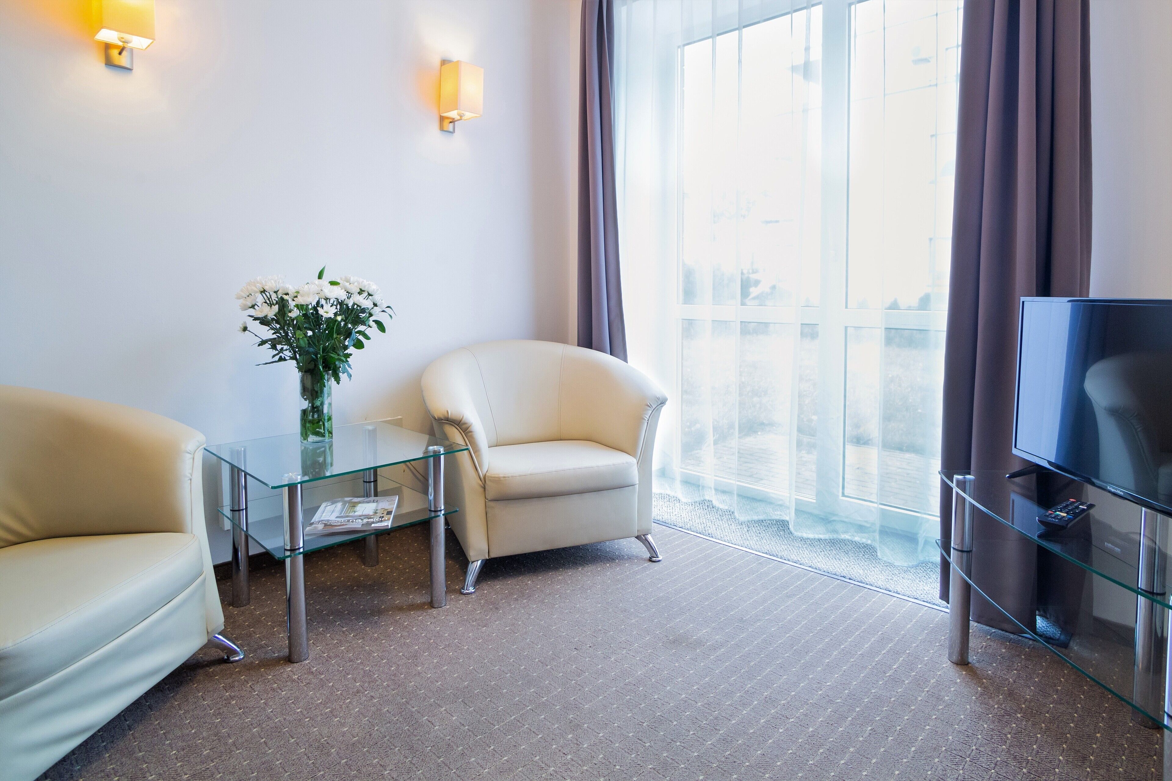 A modern hotel room with a glass coffee table, two armchairs, a flat-screen TV, and a large window with curtains.