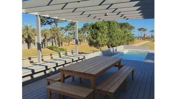 A wooden deck with benches and a pool area, surrounded by trees and a clear sky. - Best Villa in José Ignacio - Pinar del Faro - Brand New House (Departamento de Maldonado)
