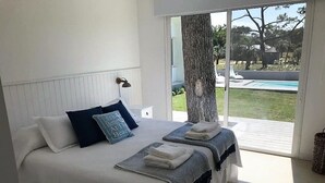 A bedroom with a large bed, two bedside tables, a window with a view of a pool and greenery, and a tree outside. - Best Villa in José Ignacio - Pinar del Faro - Brand New House (Departamento de Maldonado)