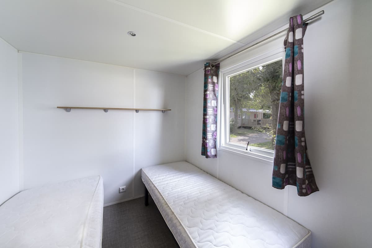 Quarto pequeno com uma cama de solteiro, janela, prateleira e cortina.