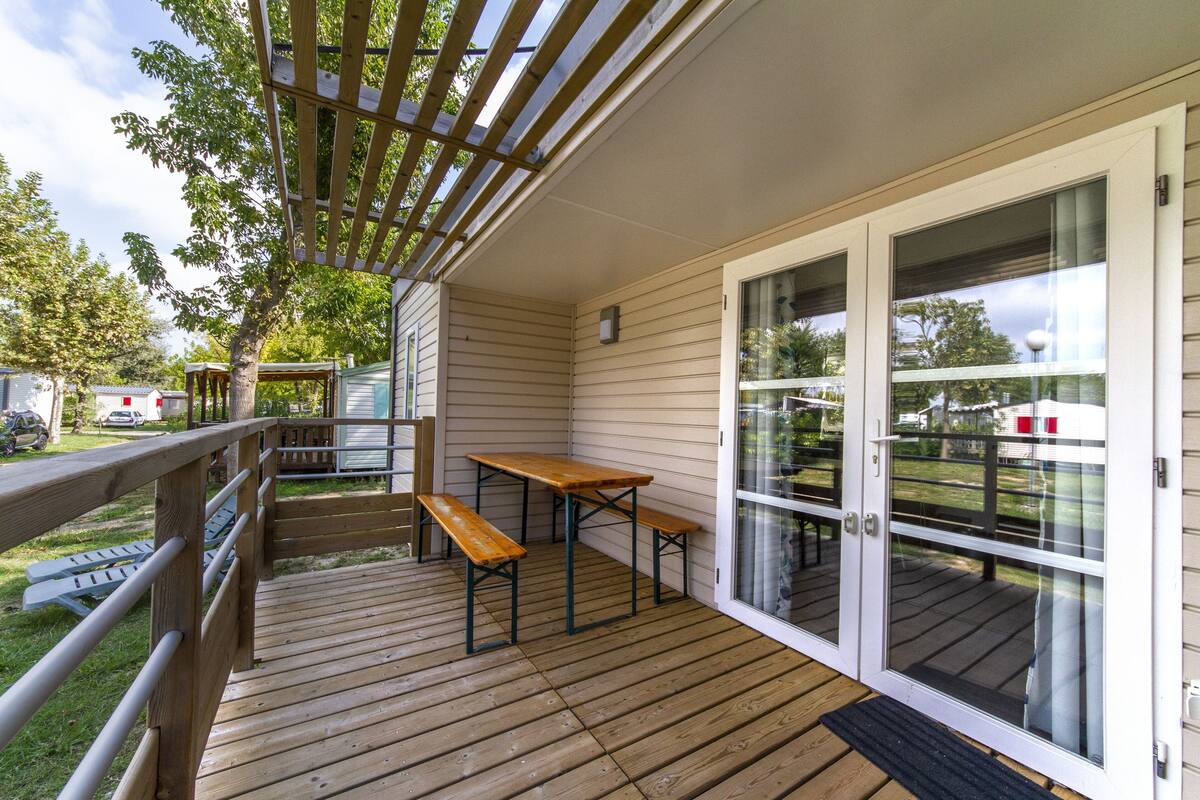 Varanda coberta com mesa e bancos de madeira, pérgola e porta de vidro deslizante que dá para um quintal.