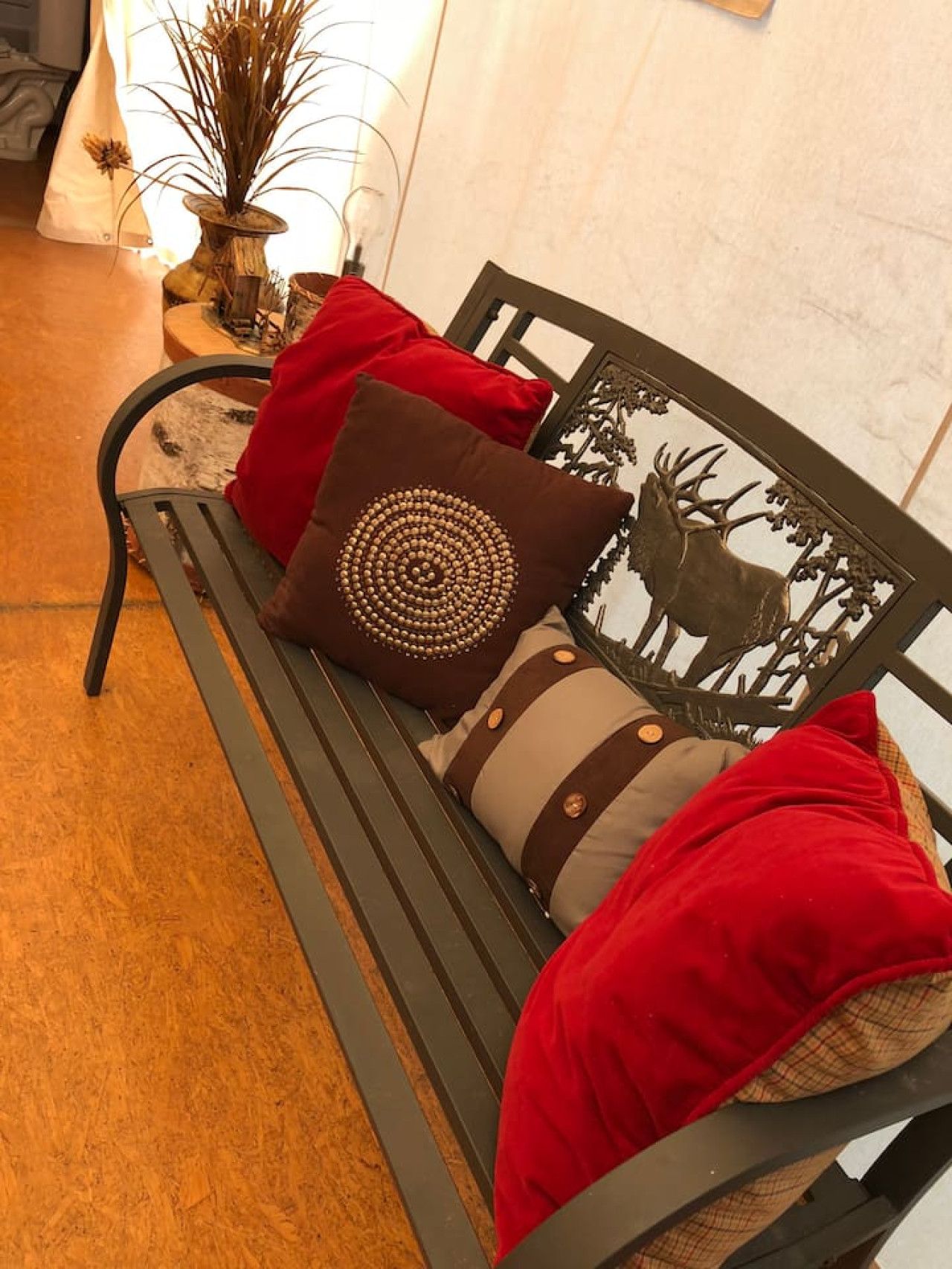 A bench with red and brown cushions, a decorative metal plaque with a deer, and a vase with dried plants.