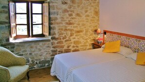 A bedroom with a stone wall, a bed with striped bedding, a chair, and a window with wooden shutters.