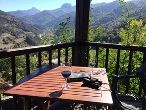 Une table en bois, sur laquelle sont posés un verre à vin et des jumelles, se trouve sur un balcon donnant sur un paysage montagneux.