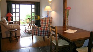 A cozy living room with a sofa, armchair, and a dining table with chairs. There is a lamp on the table and a vase with flowers.