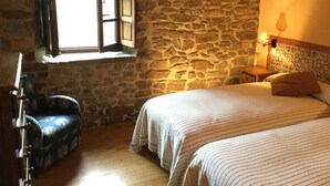 A room with stone walls, a bed with striped bedding, a chair with a floral pattern, a wooden nightstand, and a window with open shutters.