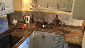A compact kitchen with white cabinets, a granite countertop, a stainless steel sink, a kettle, a bottle of wine, and a fruit bowl.