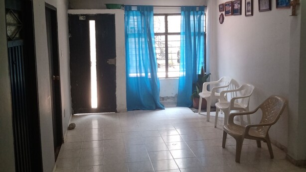 A hallway with tiled flooring, a door leading to another room, and a window with blue curtains. There are plastic chairs and a potted plant in the room. - Barranquilla residencia norte (Cali)