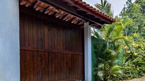 A wooden garage door with a tiled roof, a white wall, and a green fence with tropical plants. - Casa Viela da Paz (Ilhabela)