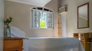 A bedroom with a bed, a nightstand, a window with shutters, a mirror, and a small wooden shelf. - Casa Viela da Paz (Ilhabela)