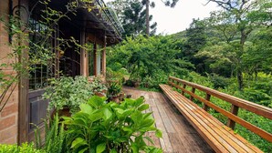 A wooden bench with a metal railing, surrounded by lush greenery and a brick building with large windows. -  Vale do Lajeado - Campos do Jordão (Campos do Jordão)