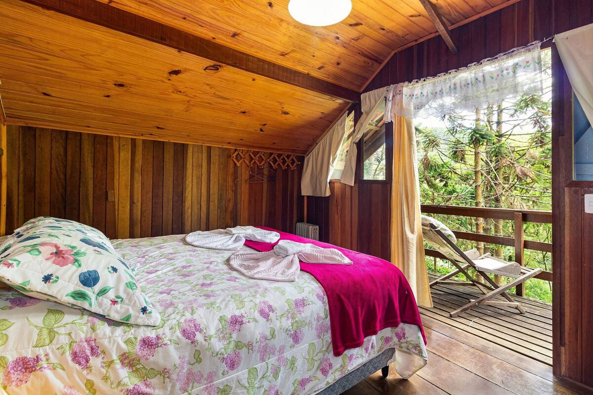 A wooden cabin bedroom with a bed, floral bedding, a red blanket, and a balcony with a chair.