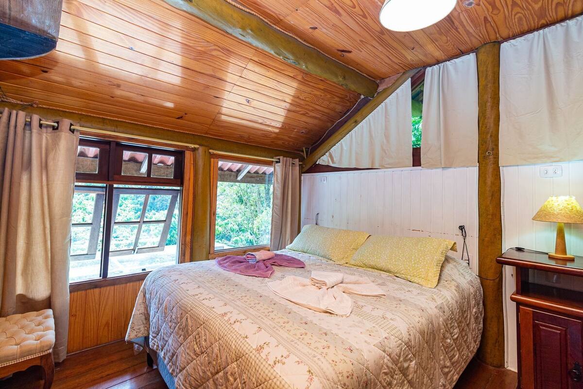 A cozy bedroom with a wooden ceiling, a bed with floral bedding, a bedside table with a lamp, and a window with curtains.