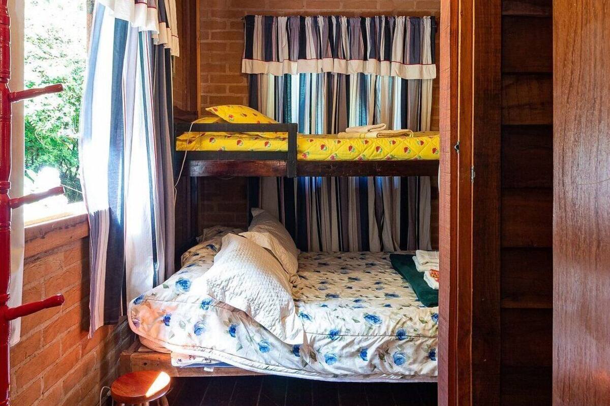 A cozy cabin room with a wooden ceiling, a bunk bed, a small stool, and a window with curtains.