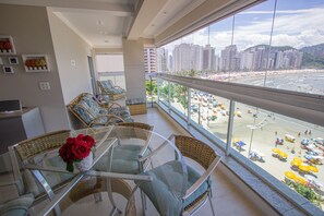 Uma varanda com móveis de vime, uma mesa de vidro com um vaso de rosas vermelhas e vista para uma praia com guarda-sóis e edifícios ao fundo.