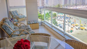 Varanda com mobiliário de vime, mesa de vidro e vista para uma praia movimentada com guarda-sóis e pessoas.