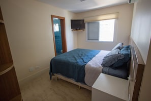Quarto com uma cama, televisão fixada na parede, janela com persianas e porta de acesso a outro cômodo.