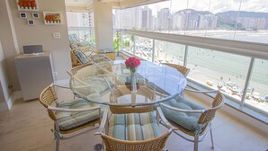 Uma varanda com cadeiras de vime e uma mesa de vidro, com vista para uma praia e edifícios altos.
