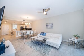 Una sala de estar moderna con un sofá blanco, una mesa de centro y un comedor con sillas, y una cocina al fondo.