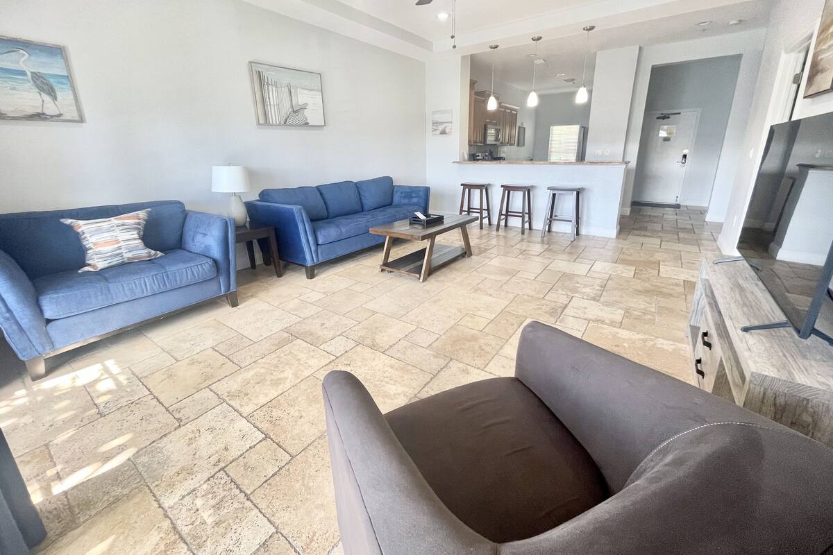 A living room with a blue sofa, a coffee table, and a grey armchair. There is a kitchen area in the background with a bar and stools.
