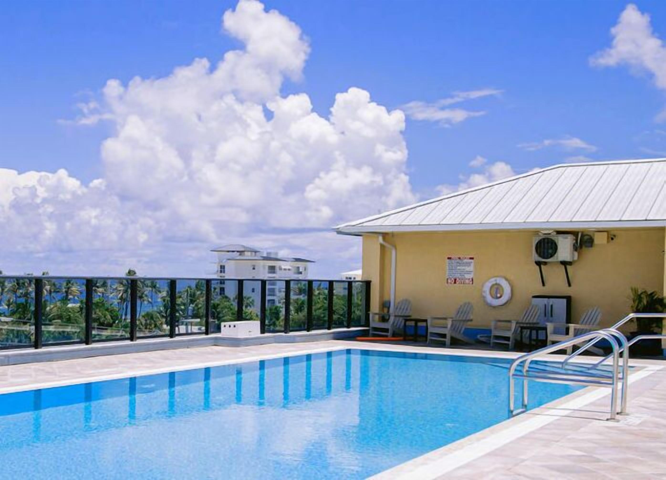 A rooftop pool with clear blue water, a yellow building with a white roof, and a view of palm trees and a distant building.